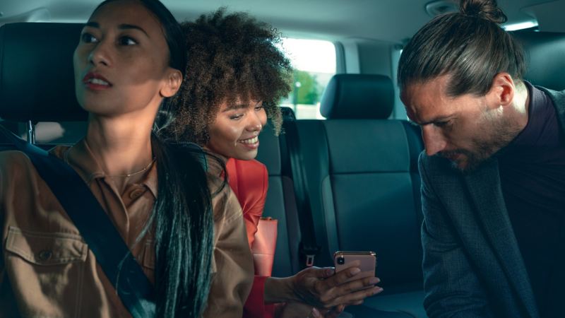 Mężczyzna i dwie kobiety siedzą w przedziale pasażerskim Volkswagena i patrzą na ekran smartfona.wo ladies are sitting the passenger compartment of a Volkswagen looking at a smartphone.