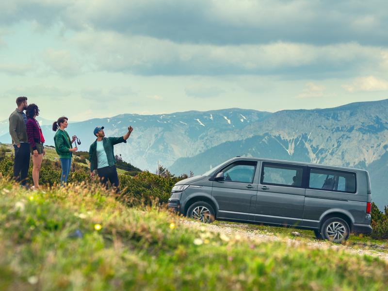 A group of people is standing in front of a Volkswagen Caravelle in the Alps.