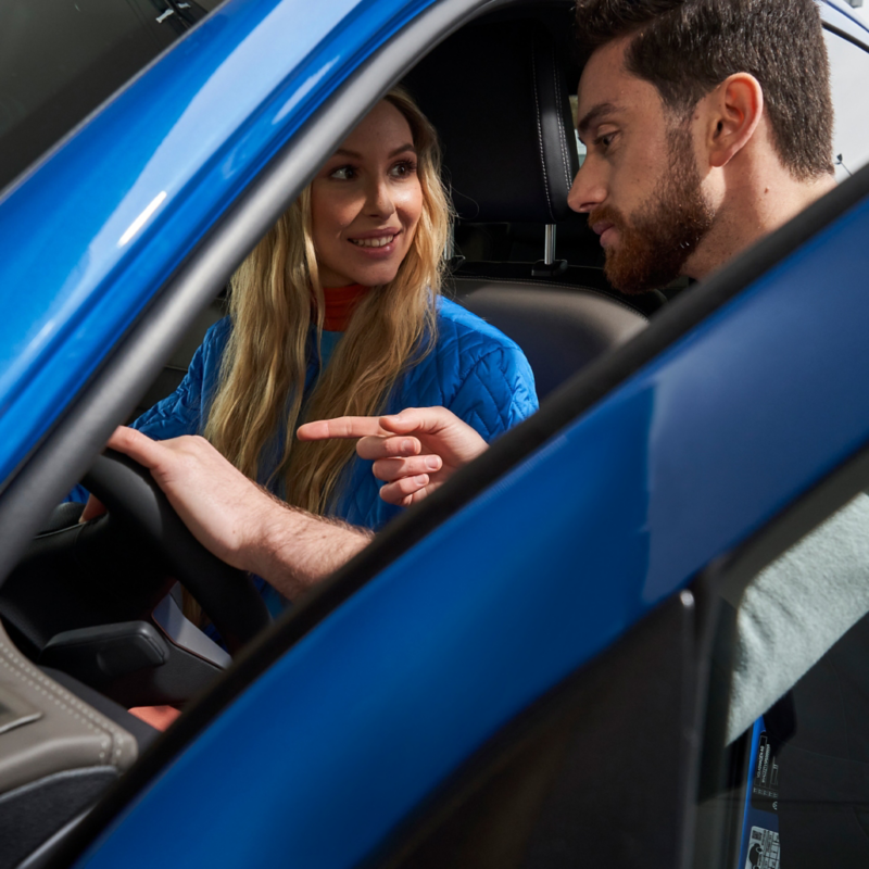 Kundin sitzt im Auto, ein Mann erklärt ihr etwas am Infotainment-System