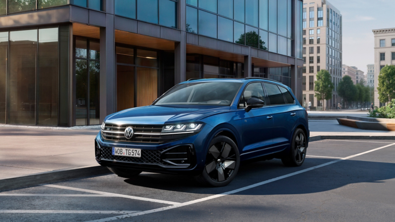 Un VW Touareg bleu est garé sur un parking devant un immeuble de bureaux moderne – vue avant élégante en environnement urbain.