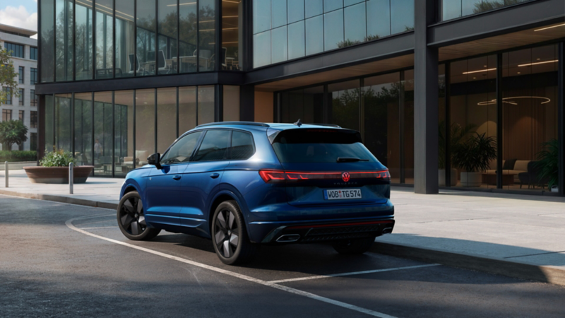 Un VW Touareg bleu de troisième génération est garé devant des façades vitrées – vue arrière nette avec bande lumineuse LED continue.