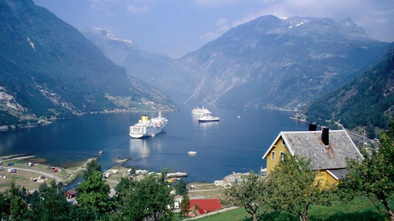 Zu sehen sind drei Schiffe in einem Fjord. Im Vordergrund ist das Festland mit Häusern erkennbar, im Hintergrund befinden sich Berge.