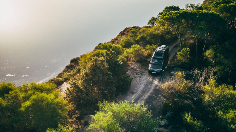 Vue d'en haut d'un Grand California vw foncé qui roule sur un chemin de terre au bord d'une falaise verdoyante. La scène, baignée dans la lumière chaude du soleil couchant, surplombe une étendue d'eau calme.