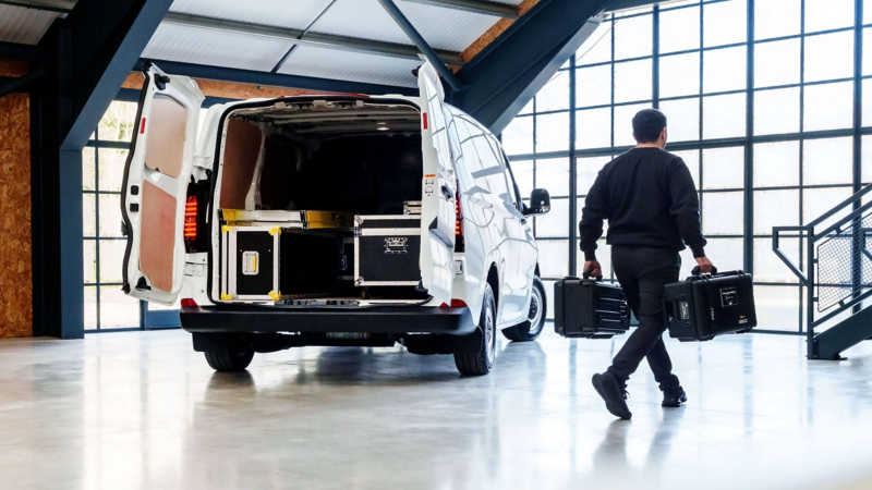 Rear view of a white VW e-Transporter Panel van with its doors opened and a man walking past it wit 2 bags.