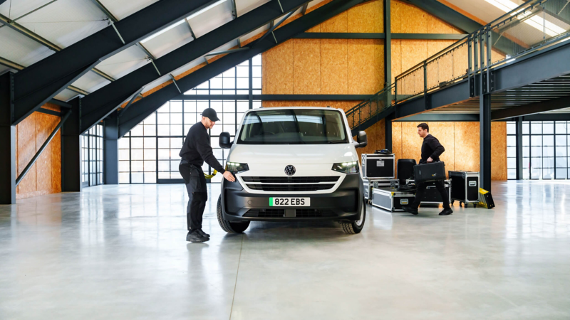 A white e-Transporter Panel van in a warehouse building with a man plugging in a charger at the front and another man loading 2 bags into the side doors