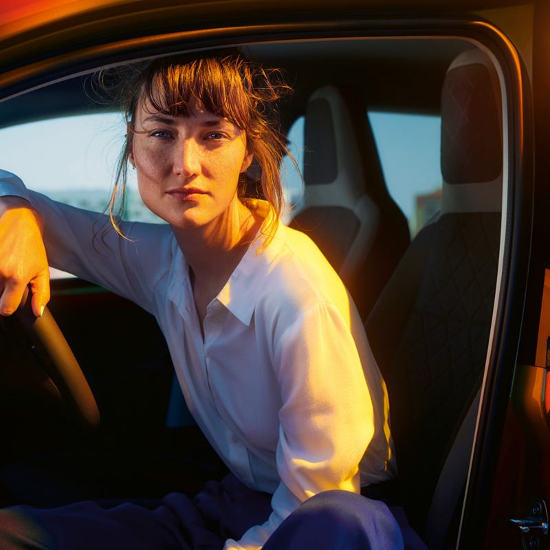Une femme est assise dans le véhicule derrière le volant d’une E-Up! de Volkswagen et regarde le coucher de soleil par la portière ouverte.lien vers la page Offres spéciales