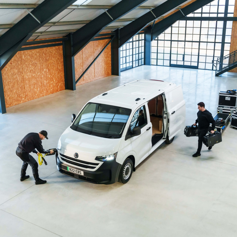 A white e-Transporter Panel van in a warehouse building with a men plugging in a charger at the front and another man loading 2 bags into the side doors