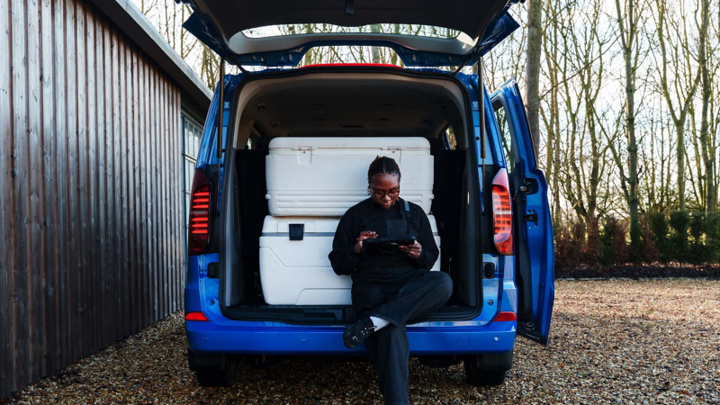 An electric VW e-Transporter van with the back doors opened with a woman sat on the bumper