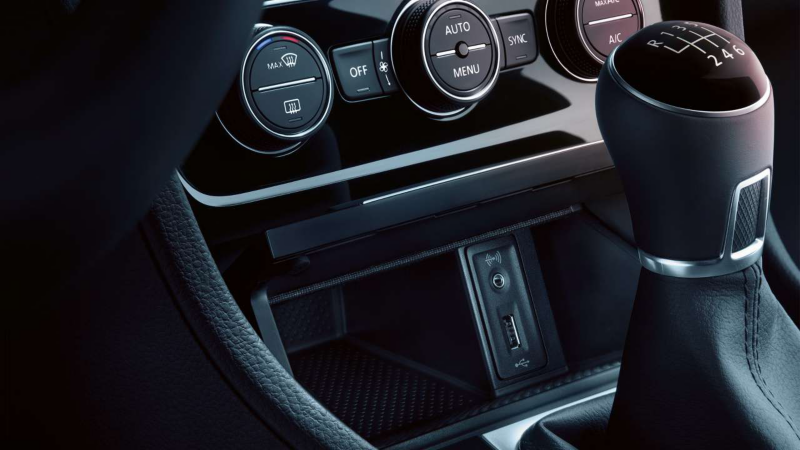 Gros plan sur l'intérieur d'une voiture VW, montrant la console centrale avec le levier de vitesse automatique, des commandes de climatisation, et un compartiment de rangement avec des ports USB et auxiliaires.