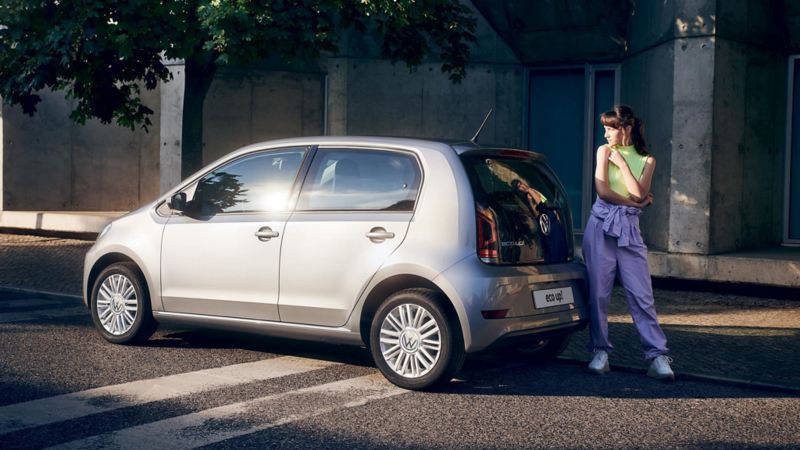Una ragazza in piedi dietro a Volkswagen eco-up!, vista 3/4 posteriormente e parcheggiata a bordo di una strada cittadina.