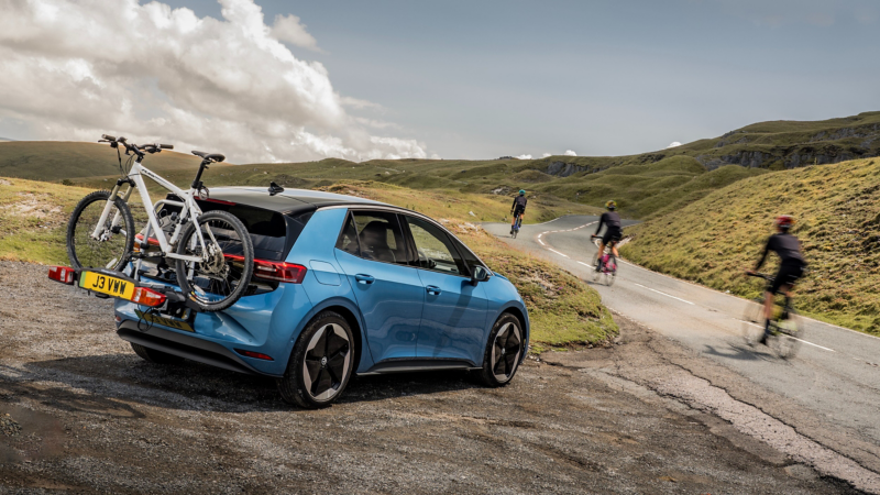 A blue Volkswagen ID.3 with a bike rack and white bicycle is parked on a scenic, winding road. Three cyclists ride past on rolling green hills under a cloudy sky.