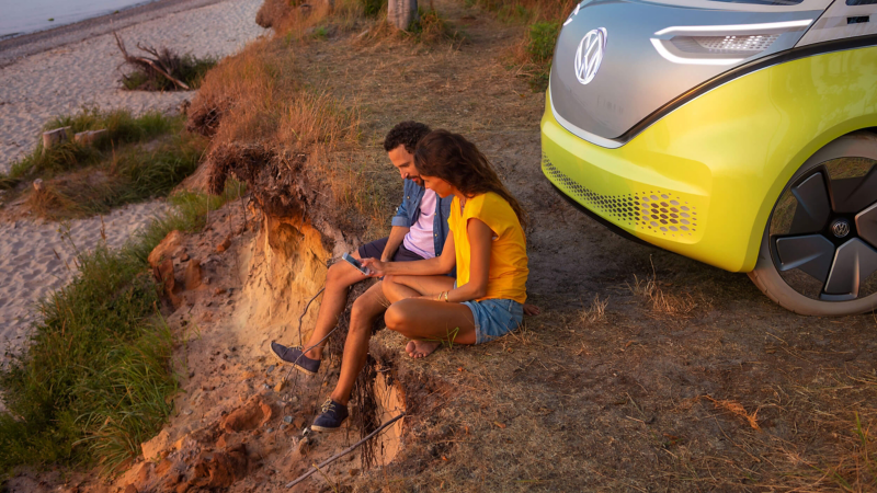A man and woman sitting by the coast beside their Volkswagen ID.Buzz