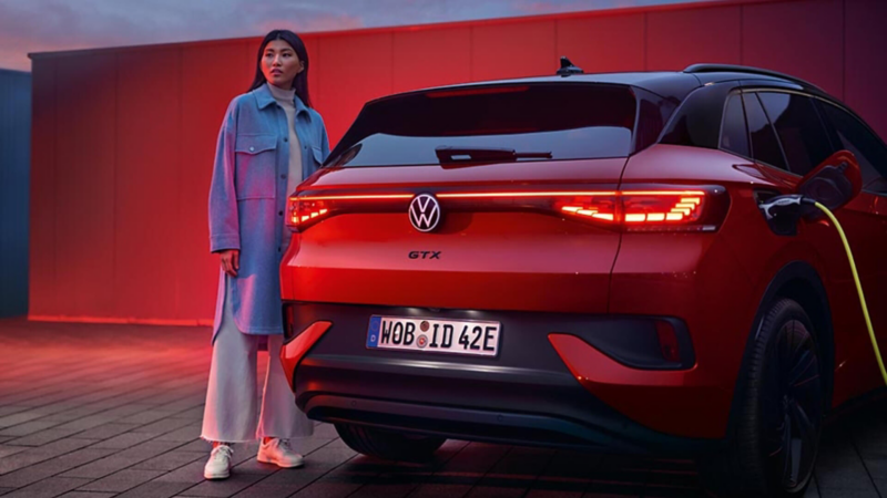 Woman in a blue coat posing next to the rear of a red Volkswagen ID.4 GTX charging at dusk.