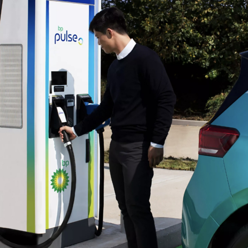 A man in a black sweater reaches for a charging cable at a bp pulse public electric vehicle charging station, with a teal car parked beside it.