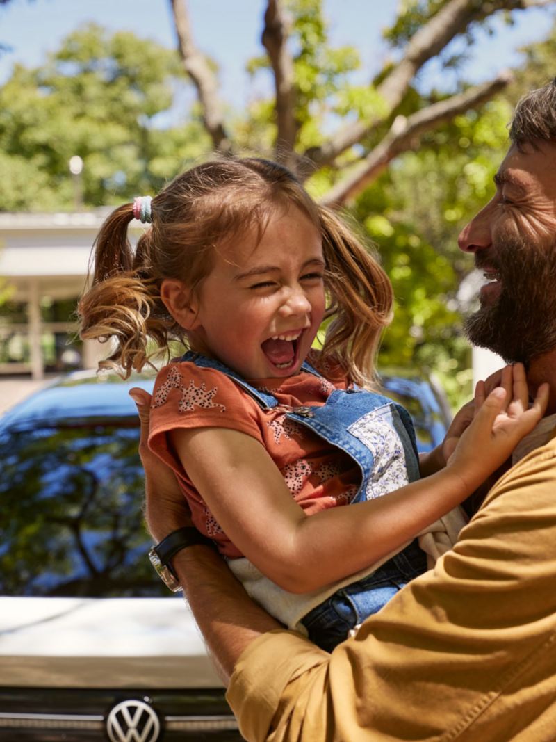 A man has a laughing child in his arms, the front of a VW ID.7 can be seen in the background.