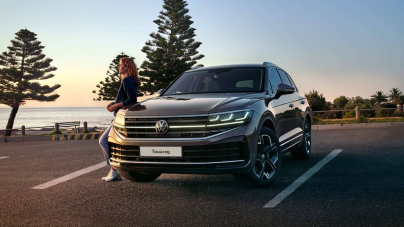 a new Touareg parked by the ocean with a woman leaning against the side of the car