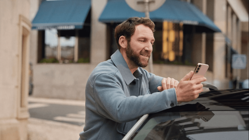 A man leaning on a car roof looking at a smartphone.