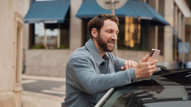 A man leaning on a car looking at the phone in his hand