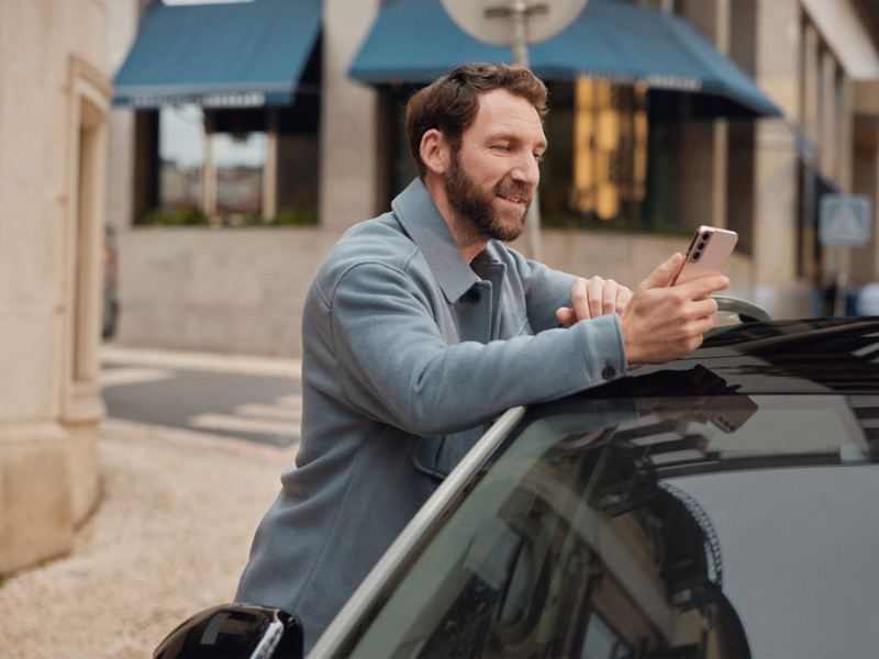 a man leaning against the back of a Volkswagen car, holding his phone in one hand and charger in the other 