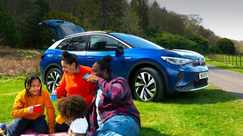 A family on a camping trip sat in front of a blue ID.4 enjoying a hot drink. The boot of the car is open.