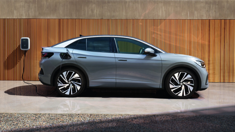Silver Volkswagen ID.4 charging at a wall charger, set against a wooden panel background. The sun casts a warm light, creating a sleek and modern feel.