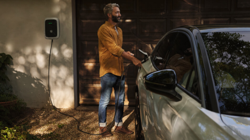 A smiling man plugs a charging cable from a wallbox charger into an electric vehicle parked in front of a garage.