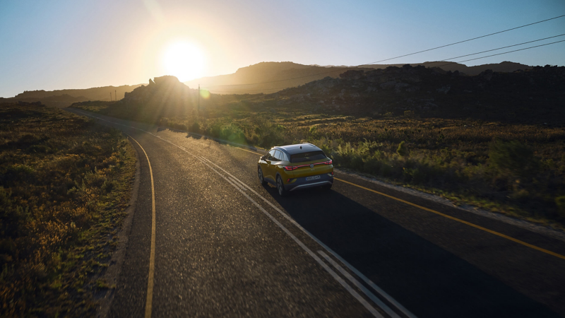 A yellow Volkswagen ID.4 drives along a winding road through a mountainous landscape at sunset. The sun casts warm light over the scenery, creating a serene atmosphere.