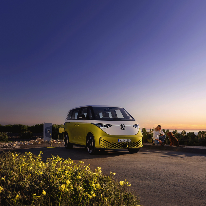 At dusk on the beach, a woman is petting a dog, a VW ID. Buzz is parked next to it.