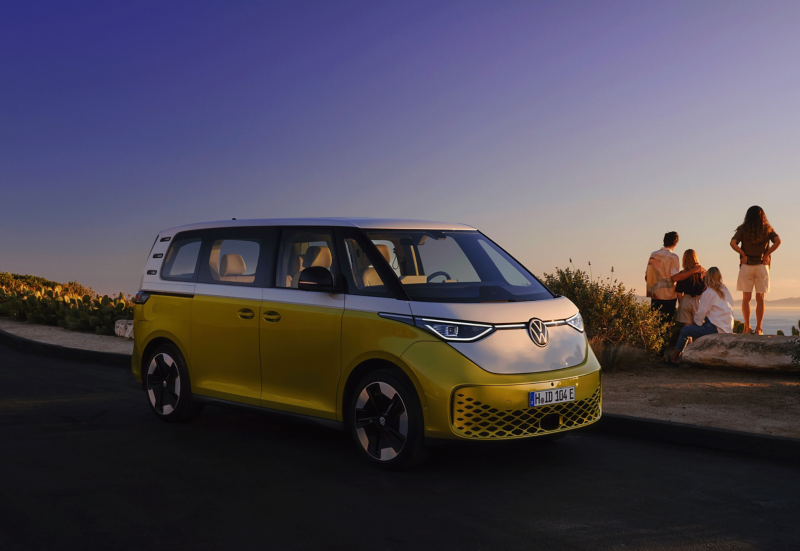 A yellow and white VW ID. Buzz stands to one side on the edge of a beach promenade. In the background, a family with two children looks into the sunset over the sea. 