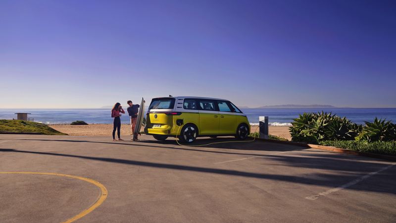 The VW ID. Buzz parks on the beach, in front of it a man shows his daughter a surfboard.