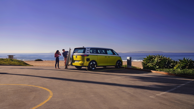 The VW ID. Buzz parks on the beach, in front of it a man shows his daughter a surfboard.