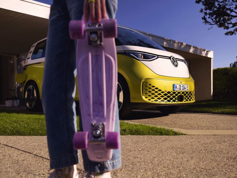The ID. Buzz stands in a driveway, with a girl holding a purple skateboard in the foreground. 