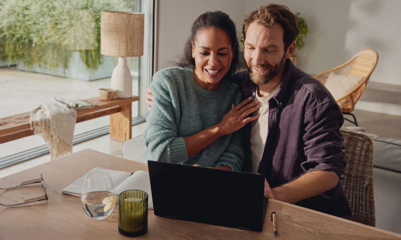 A smiling couple sitting together by a table and using a laptop
