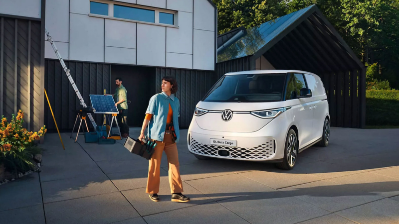 A white Volkswagen ID. Buzz Cargo van is parked outside a modern building with dark paneling and a sloped roof. A woman in a light blue work shirt and brown trousers walks past carrying a toolbox, looking to the side. In the background, a man in a green shirt is setting up solar panels with a ladder and equipment nearby. The scene is illuminated by warm sunlight, with greenery and flowers visible on the left.