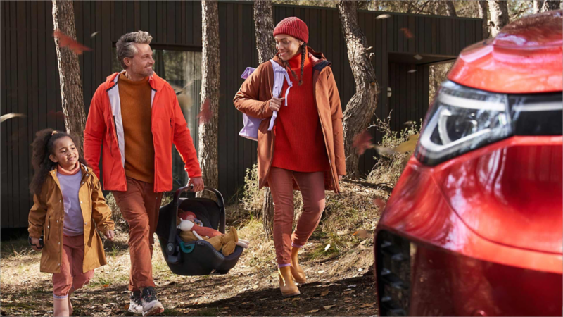 Un hombre sujetando un bebe en una sillita de coche junto a una niña y una mujer al lao de un Volkswagen rojo