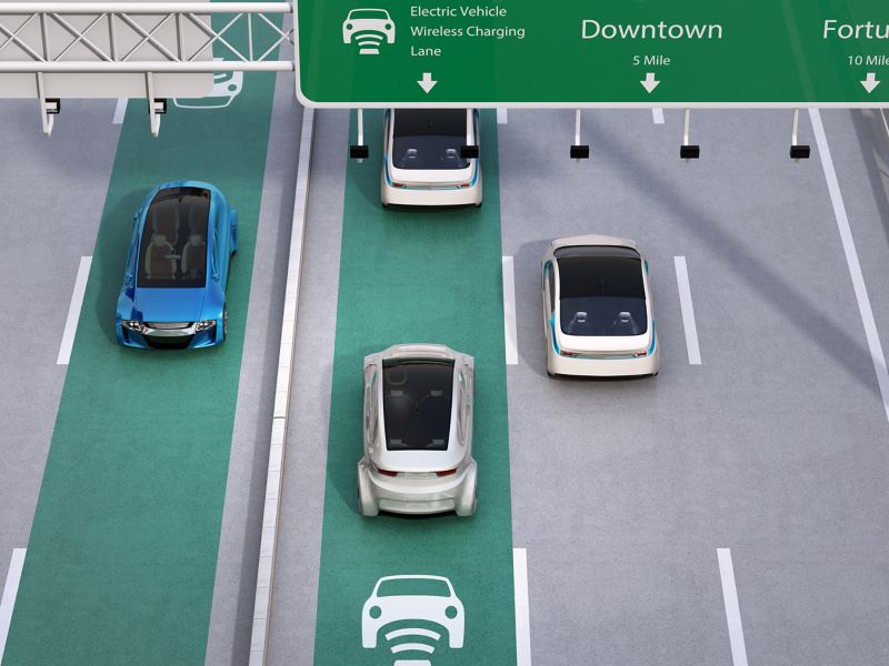 Elektroautos fahren auf einer grün und mit Sinnbild gekennzeichnete Fahrbahn auf einer mehrspurigen Straße, auf der induktives Laden möglich ist.