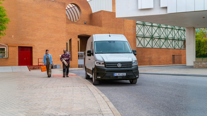 Una furgoneta blanca de Volkswagen aparcada en un exterior citadino con dos personas trabajadoras cerca