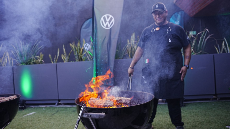 Chef con gorra y delantal prepara carne en una parrilla con llamas, junto a un panel de Volkswagen.