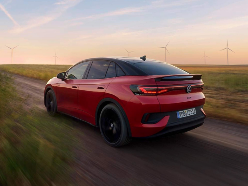 Rear three-quarter view of a red Volkswagen ID.5 GTX electric SUV driving on a dirt road with wind turbines in the background.