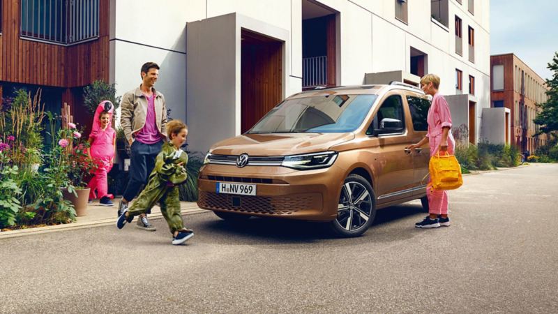 Véhicule utilitaire Volkswagen Caddy brun stationné devant une maison. Une femme ouvre la portière conducteur et un enfant avance devant le véhicule.