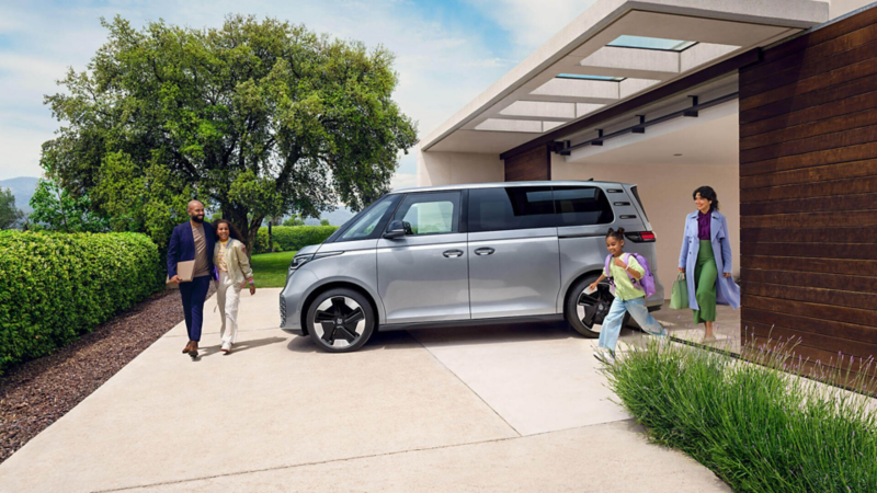 Véhicule utilitaire Volkswagen ID. Buzz gris métallisé stationné dans une entrée de garage avec une famille de part et d'autre de la voiture. 