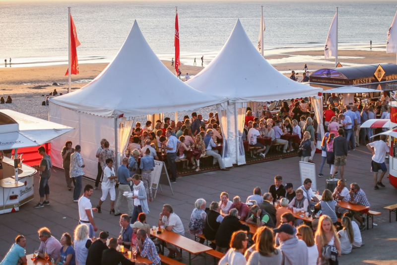 På stranden er der opstillet hvide telte og ølbænke: Folk stimler sammen i forbindelse med en surfkonkurrence.