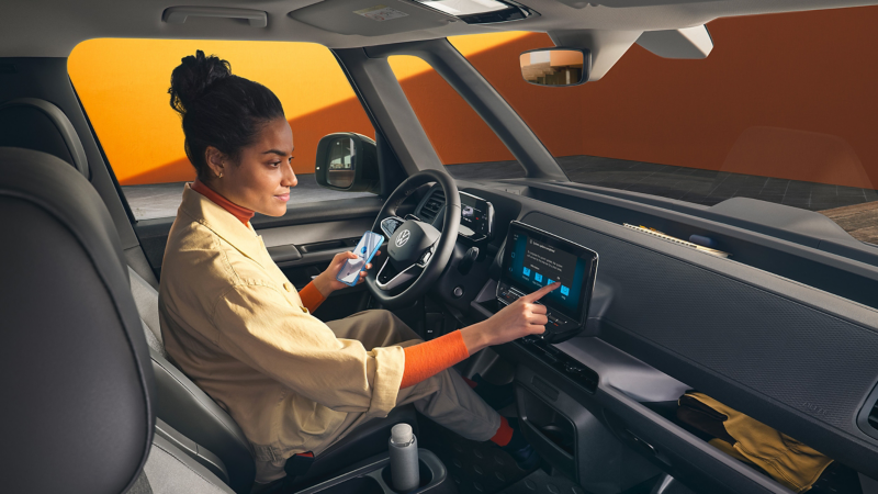 Woman sitting in the driver’s seat of a Volkswagen van, using the touchscreen infotainment system while holding a smartphone.