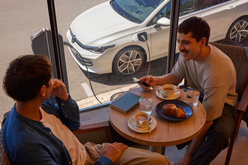 Deux homme sont assis dans un café, une VW Golf eHybrid en charge est visible par la vitrine.