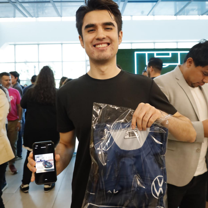 Joven sonríe mostrando playera de color azul de Volkswagen y pantalla de celular.