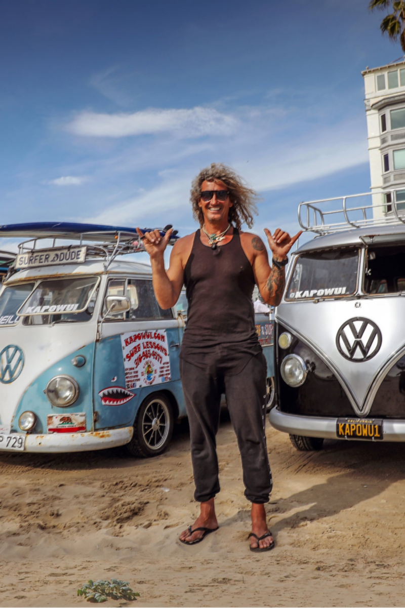 Surferen Martin står på stranden foran to gamle folkevognsrugbrød. Han laver en ”Hang Loose“-gestus. 