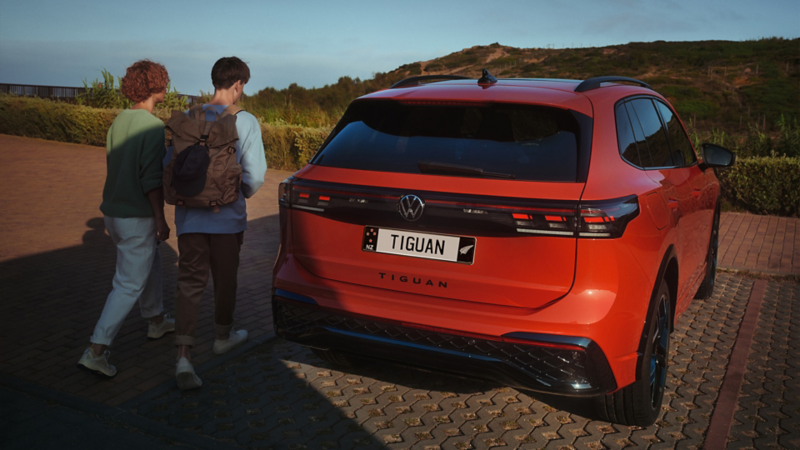 A young man on the driver side of the red VW Tiguan operates the optional Park Assist Plus from his smartphone.