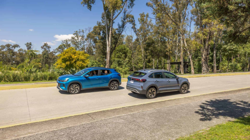 Dos camionetas Volkswagen de colores azul y gris avanzan por avenida rodeada por árboles. 