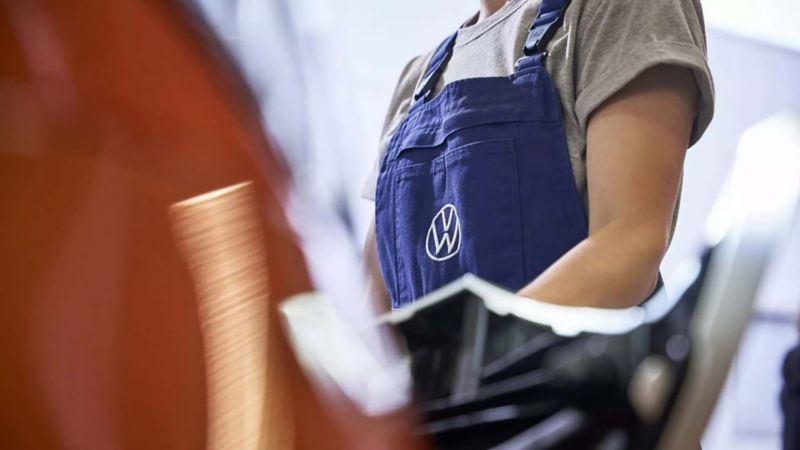 Técnica de Volkswagen usa overol azul con logo VW durante trabajo dentro de taller mecánico.