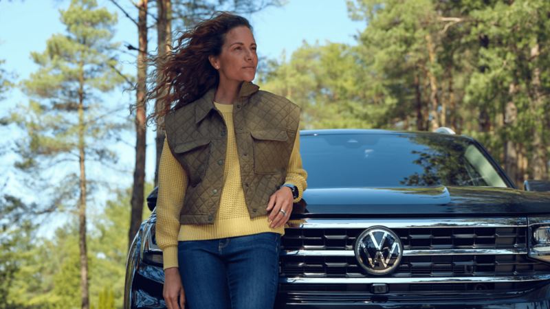 Une femme est appuyée sur le capot d'un Atlas 2022 de Volkswagen bleu tourmaline métallisé dans une forêt.